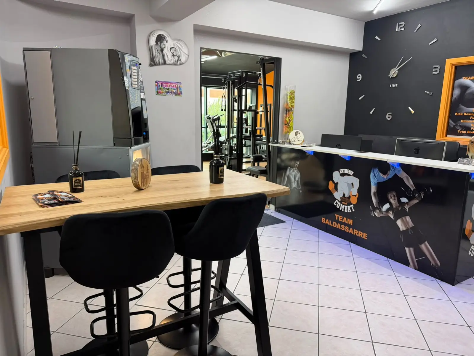 La reception con il bancone Team Baldassarre, il tavolo alto con gli sgabelli e la sala pesi sullo sfondo